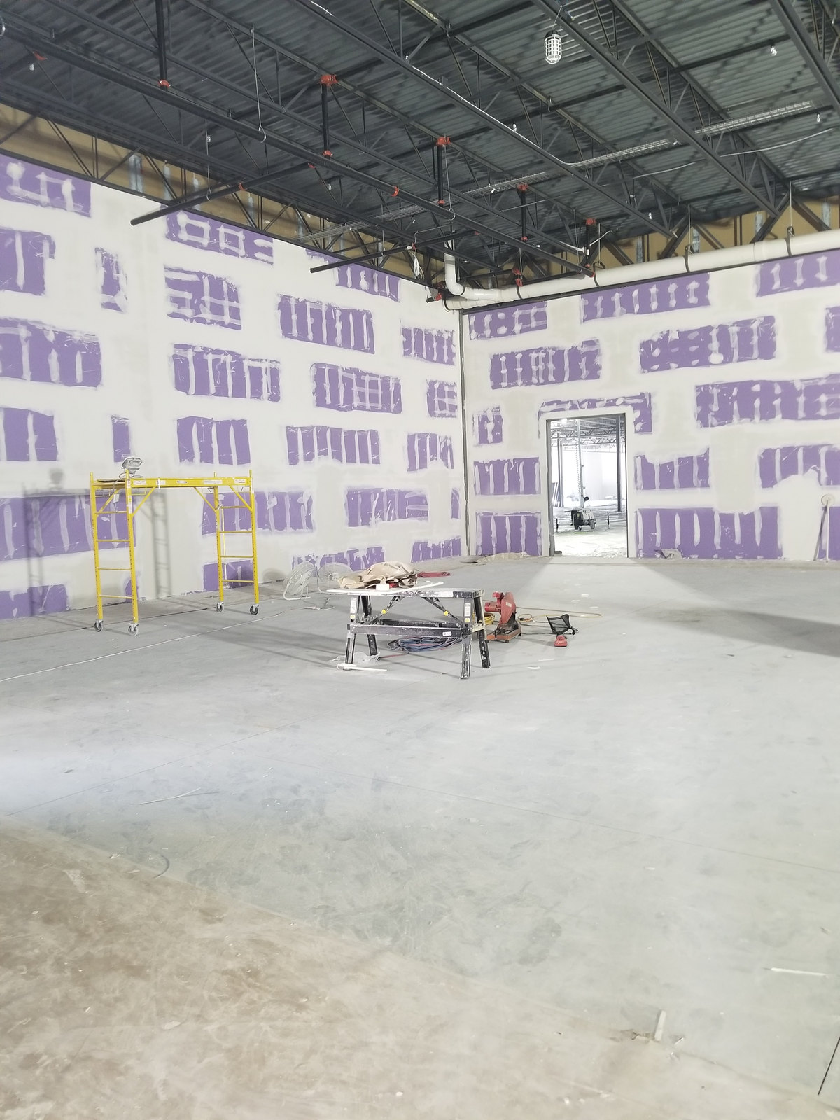 Large interior with purple wall panels, white joint treatment, exposed roof structure and a yellow scaffold.