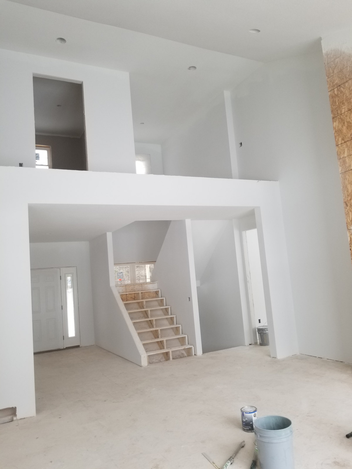 Double-height white interior with large openings, a stairway and recessed-light openings during construction.