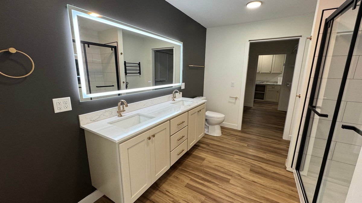 Bathroom with a double vanity, illuminated rectangular mirror, dark accent wall and glass shower enclosure.