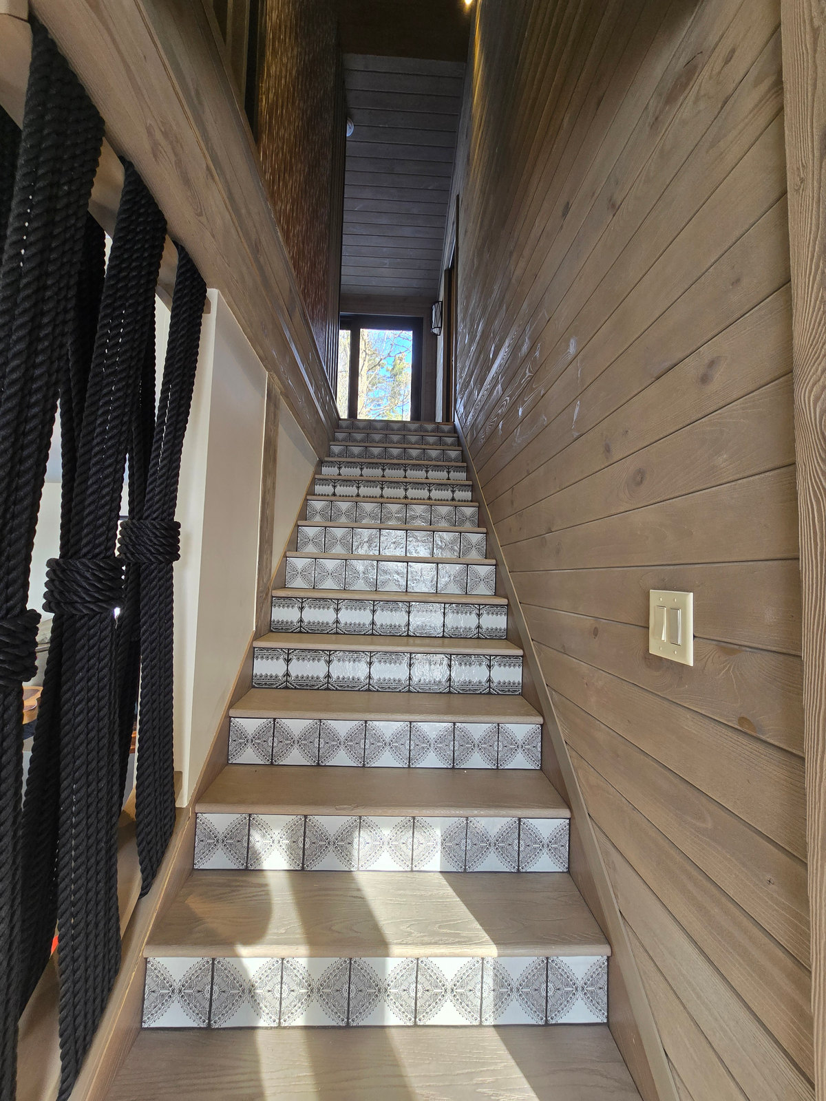 A staircase with patterned risers, pale wood-clad walls and a window at the top.