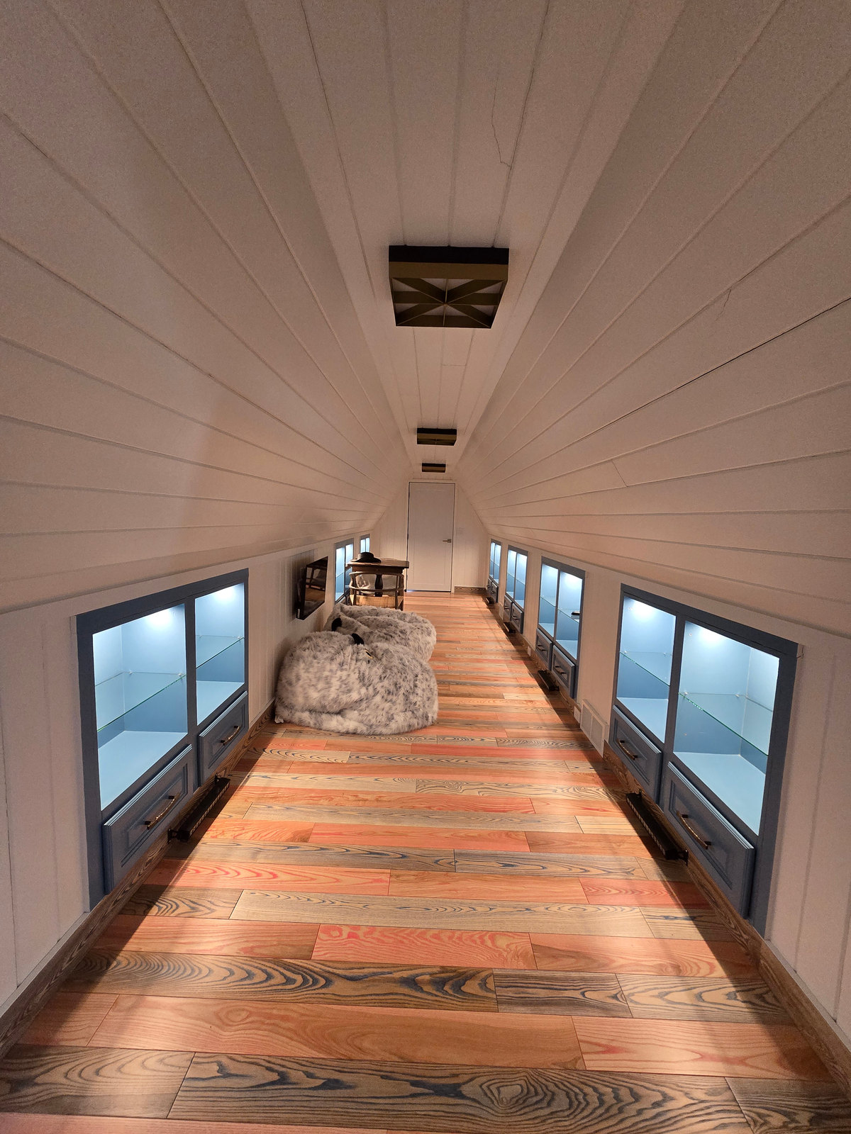 A long passage with pale sloping plank walls, illuminated display cabinets and a multicolored wood floor.